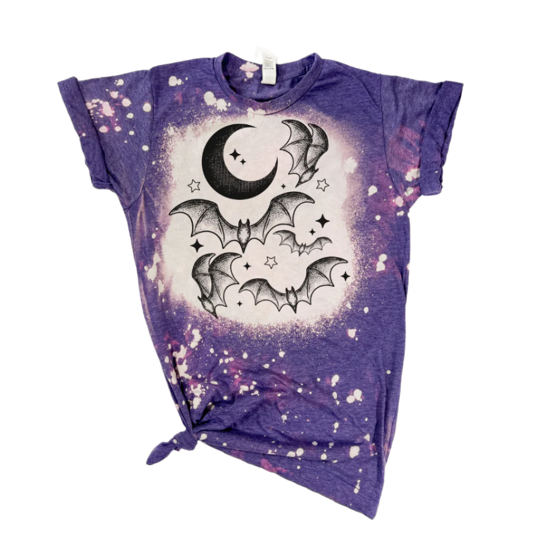 A purple bleached t-shirt with a graphic of a crescent moon and bats, and white splatters around the design.